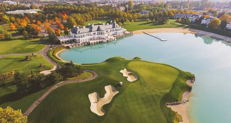 Vue aérienne d'un complexe de golf au bord du lac avec un club-house majestueux, une plage de sable blanc et de vastes fairways bordés d'arbres d'automne.