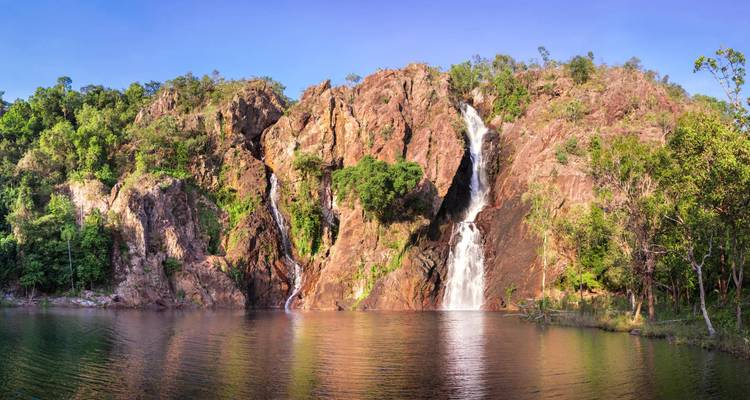 Twin waterfalls cascade down red rock cliffs into a tranquil plunge pool surrounded by forest.