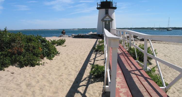 Petit phare blanc situé à côté d'une plage de sable fin et d'une promenade en bois offrant une vue sur les eaux bleues de l'océan.