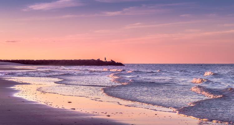 Un coucher de soleil pastel serein sur un rivage sablonneux tranquille avec des vagues douces reflétant des ciels roses et violets.