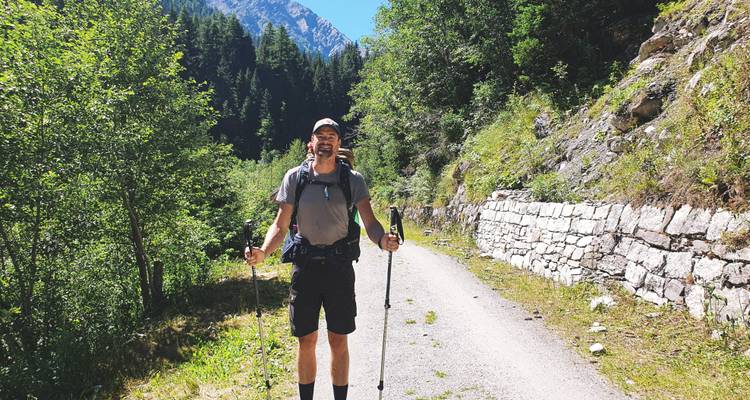 Randonneur souriant posant avec des bâtons sur un sentier de montagne boisé bordé de murs de pierre