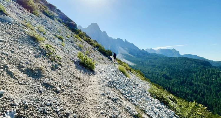 Randonneur solitaire gravissant un sentier de gravier lumineux avec des silhouettes de montagnes déchiquetées devant