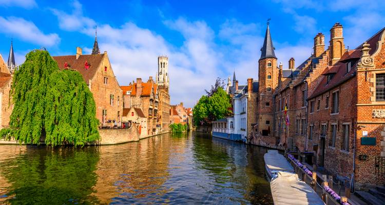 Zonnig uitzicht langs het kanaal van Brugge richting het Belfort, met bakstenen huizen en bladrijke oevers langs het water.