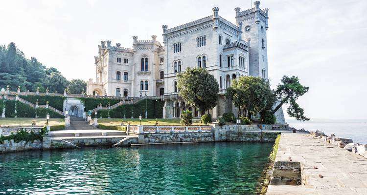 Elegant wit Kasteel Miramare gelegen op een rotsachtige kaap boven smaragdgroene kustwateren met verzorgde tuinen en sierlijke trappen.