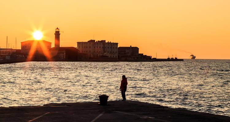 Silhouet van een persoon die op een pier staat en kijkt naar de zon die ondergaat achter een vuurtoren en industriële waterkant op kalme zeeën.