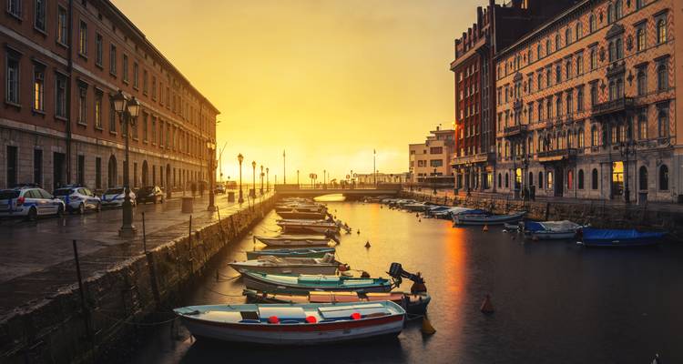 Gouden zonsopgang boven een kanaal omzoomd met kleine bootjes en historische gebouwen in Triëst, reflecties glinsterend op het water.