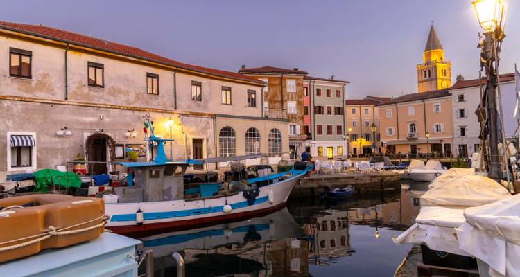 Schemerzicht van de haven van Piran met vissersboten aangemeerd naast pastelkleurige gebouwen en een hoge kerktoren onder paarse luchten.
