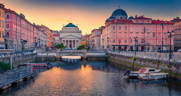 Levendige zonsondergang boven het Canal Grande van Triëst met kleurrijke gevels en de Sant'Antonio kerk weerkaatsend in het kalme water.