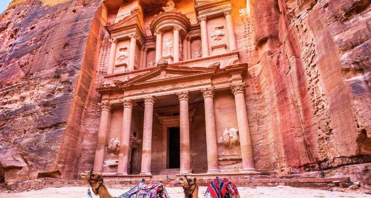 Iconische schatkistgevel van Petra omlijst door rozerode kliffen met rustende kamelen op de voorgrond.