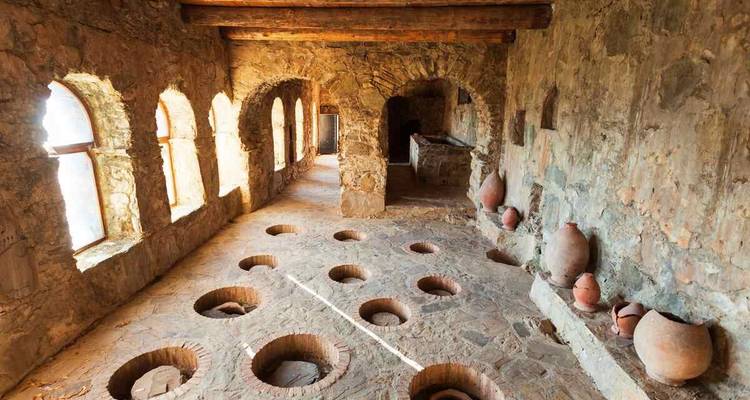 Pasillo iluminado por el sol de una bodega antigua que muestra filas de jarras de barro y pozos profundos de qvevri.