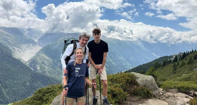 Famille de trois randonneurs à un point de vue panoramique avec des montagnes enneigées et des nuages.