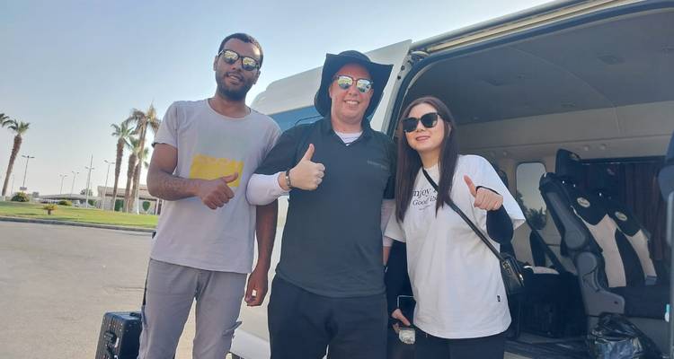 Trois voyageurs font un pouce en l'air à côté d'un van de tourisme blanc sous un ciel dégagé avec des palmiers.