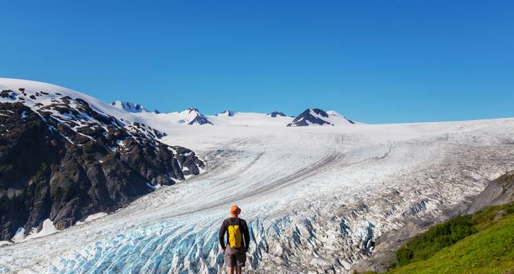 Randonneur solitaire contemple le bord d'un glacier tentaculaire sous un ciel bleu clair et des sommets enneigés