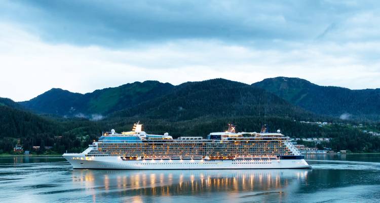 Un grand navire de croisière blanc navigue dans des eaux calmes avec en arrière-plan des montagnes boisées sous un ciel nuageux du soir.