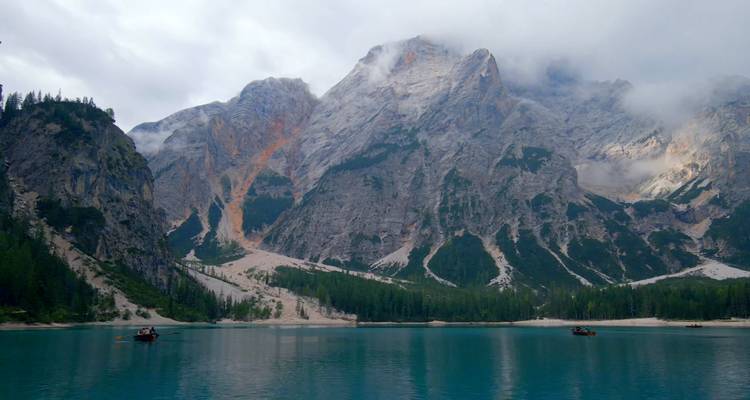 Lac turquoise avec des pics brumeux et des bateaux éparpillés sous des nuages dramatiques.