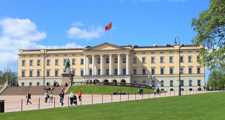 Palais Royal néoclassique d'Oslo avec drapeau flottant, entouré de pelouses vertes et de visiteurs se promenant.