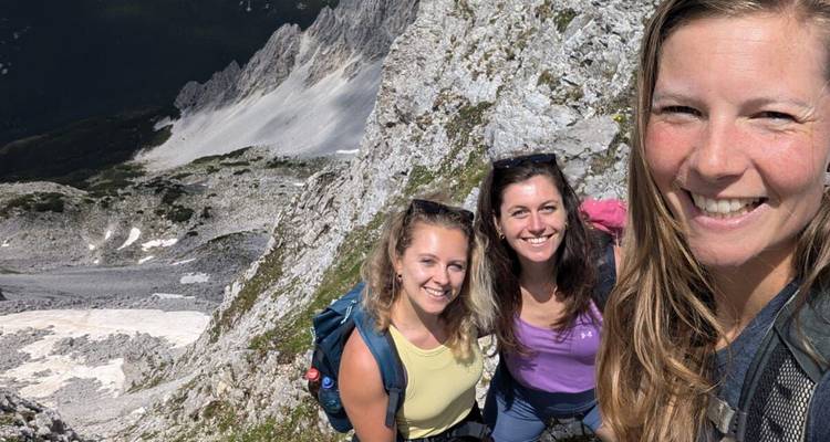 Trois femmes prenant un selfie sur un rebord étroit en hauteur au-dessus d'une vallée alpine.