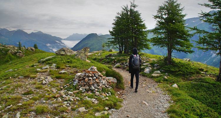 Excursionista solitario con mochila caminando por un sendero alpino pedregoso entre laderas verdes ondulantes y picos brumosos distantes.