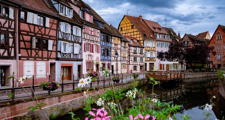 Fachwerkhäuser in Pastellfarben säumen einen friedlichen, mit Blumen geschmückten Kanal im Elsass.