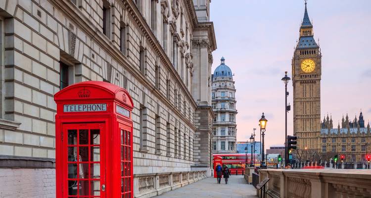 Iconische rode telefooncel naast Westminsters stenen gevel met Big Ben en een dubbeldekkerbus in de schemering.