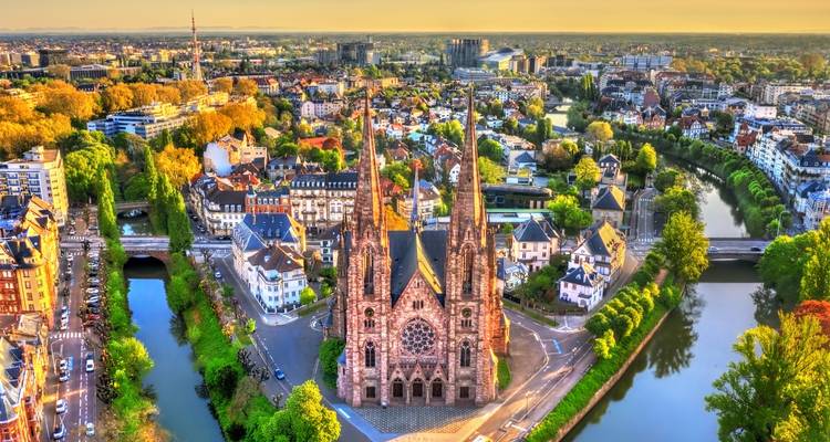 Aerial golden-hour view over Strasbourg highlighting the twin-spired Saint Paul’s Church surrounded by canals and colorful city blocks.