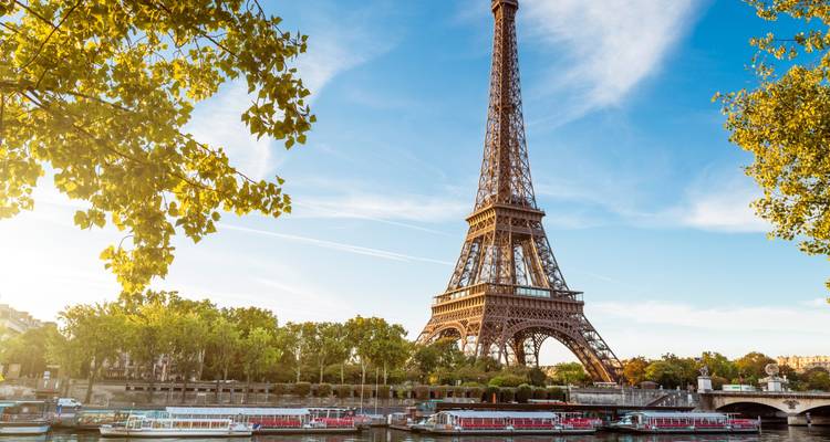 The Eiffel Tower soars above the Seine with sightseeing boats and leafy trees on a sunny afternoon.