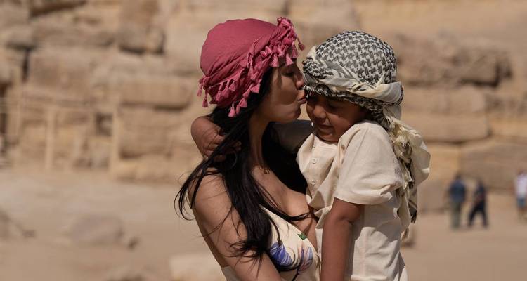Une femme embrasse tendrement un petit enfant portant un keffieh, des ruines du désert floues derrière eux.