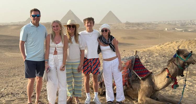 Cinq voyageurs se tiennent avec un chameau décoré sur une crête sableuse avec des pyramides dans une lumière brumeuse.