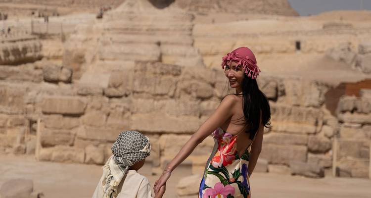 Femme en robe fleurie guide un enfant à travers des ruines antiques sous le regard vigilant du Sphinx.
