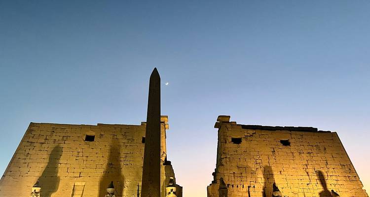 Vue crépusculaire de l'entrée du temple de Louxor avec la silhouette de l'obélisque et le croissant de lune au-dessus.