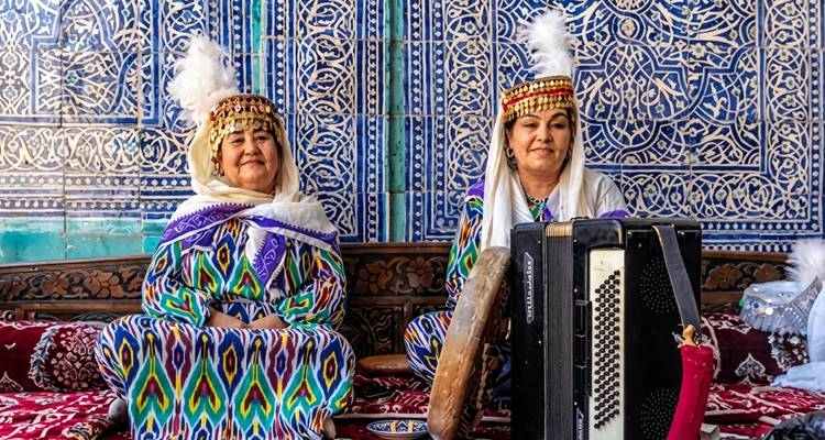 Dos mujeres uzbekas con túnicas ikat coloridas tocan con tambor y acordeón frente a azulejos azules ornamentados.