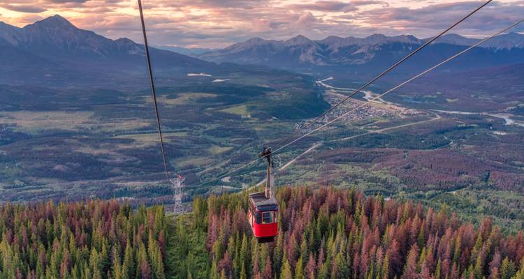 Red gondola of a mountain cable car gliding above a vast forested valley with rugged peaks at sunset.