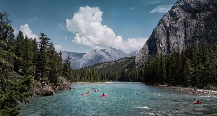 Kayakers paddling down a turquoise river flanked by dense evergreen forest and towering rocky mountains under a blue sky with clouds
