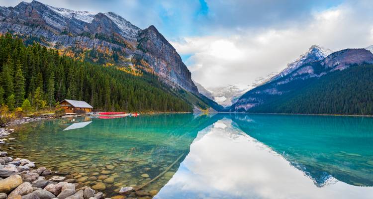 Crystal-clear Lake Louise with mirror reflection of snow-dusted peaks, red canoes and a small wooden cabin along the forested shore