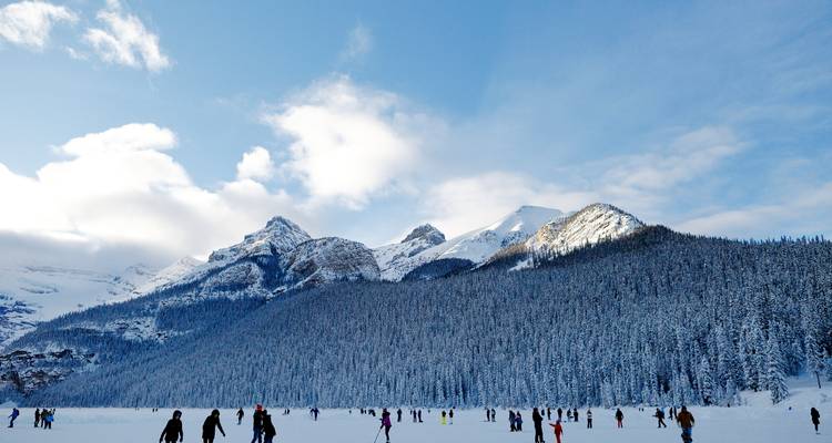 Dozens of visitors walk and skate on a frozen lake surrounded by snow-covered pine forest and craggy mountain peaks
