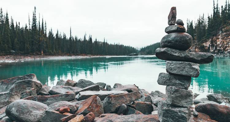 Stone cairn balanced on rocky shoreline beside turquoise glacial lake with dense fir trees in the background