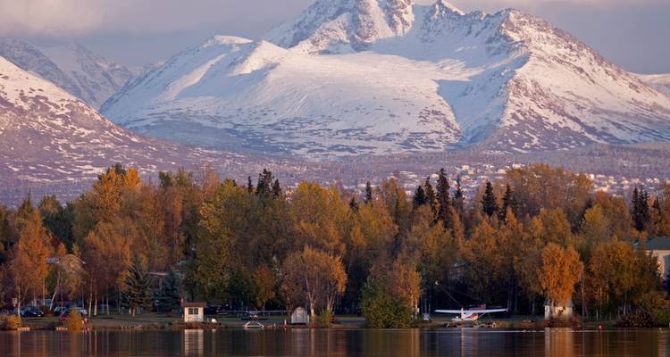 Autumn trees and floatplanes line a calm lake with snow-capped Alaskan mountains rising in the background