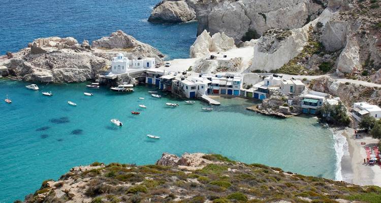 Una cala pintoresca con agua turquesa, casas de botes cícladas blancas y pequeños botes de pesca amarrados bajo acantilados escarpados.