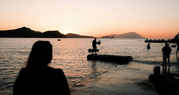 Figuras en silueta se alzan junto a las tranquilas aguas del mar al atardecer con colinas oscuras e islas en la distancia.