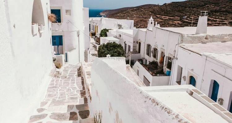 Callejón estrecho de piedra bordeado de casas cicládicas encaladas y contraventanas azules con vista al lejano mar Egeo.
