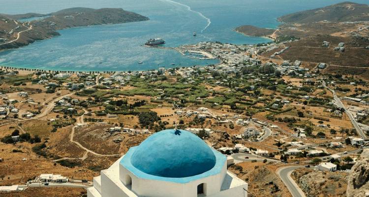 Capilla blanca de cúpula azul encaramada sobre una vista panorámica de un puerto, tierras de cultivo y costa escarpada.