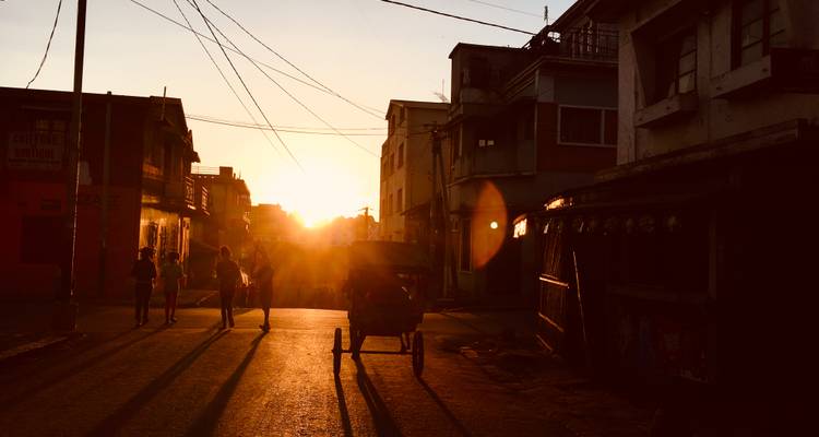 Silhouettes de piétons et d'un pousse-pousse tiré à la main dans une rue poussiéreuse au coucher du soleil.