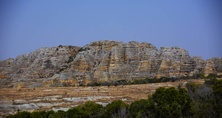 Mesa rocheuse aride avec des falaises de grès stratifiées s'élevant au-dessus de prairies sèches sous un ciel bleu dégagé.