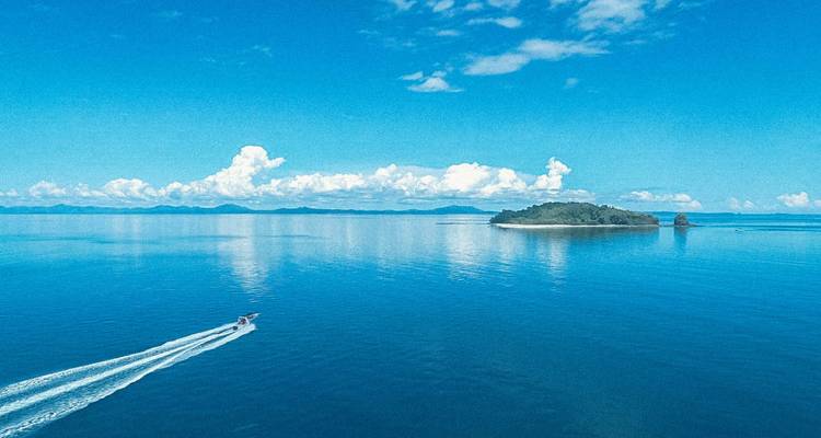 Un hors-bord laisse un sillage blanc à travers une mer turquoise infinie vers une petite île verte sous un ciel dégagé.