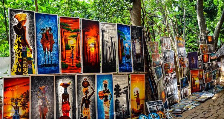 Étal de marché en plein air exposant de vives peintures africaines de faune et de culture sous des arbres feuillus.