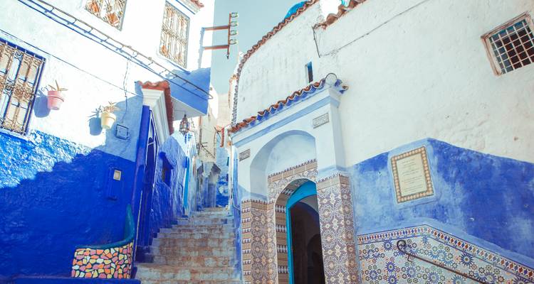 Callejón estrecho de azul vívido en Chefchaouen con entrada de azulejos y escalones de piedra empinados.