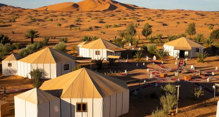 Campement de luxe dans le désert avec des tentes en toile installées parmi les dunes dorées du Sahara sous un ciel de fin d'après-midi.
