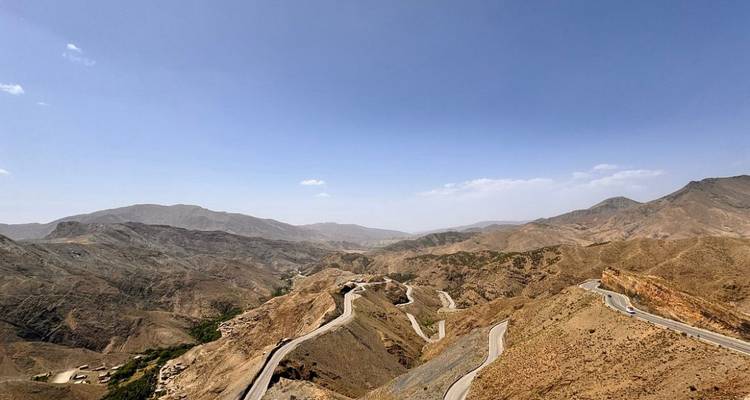 Une route de montagne sinueuse serpente à travers des pics bruns escarpés sous un ciel marocain bleu et dégagé.