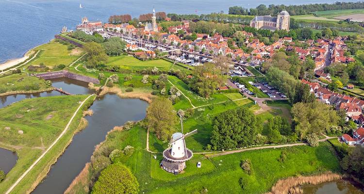 Vue aérienne d'un charmant village côtier hollandais avec un moulin à vent blanc, des maisons aux toits rouges, des voies navigables et des champs verts sous un ciel bleu éclatant.