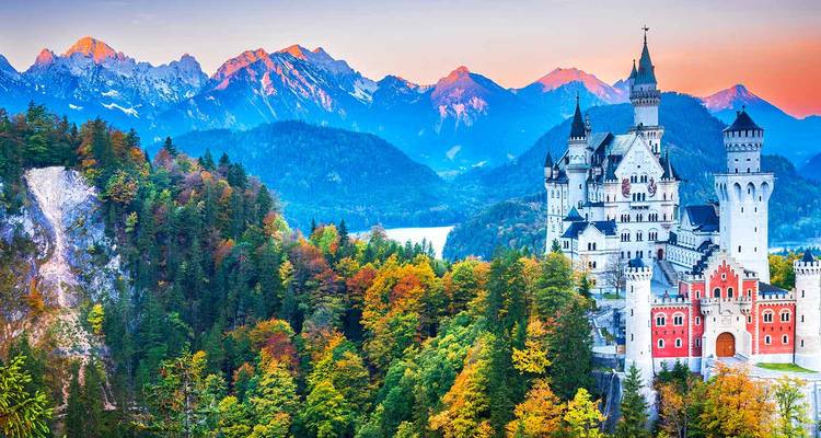 El icónico Castillo de Neuschwanstein elevándose sobre coloridos bosques otoñales con picos alpinos cubiertos de nieve al fondo durante la puesta de sol.